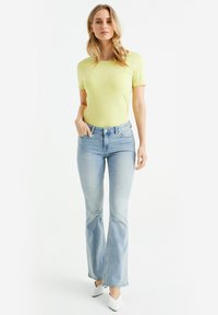Top giallo a maniche corte abbinato a jeans a zampa d'elefante di un blu chiaro. Tacchi bianchi a punta completano l'outfit.