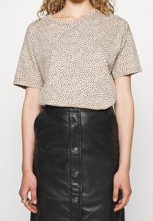 Kvinde iført beige kortærmet top med sorte pletter, stoppet ind i sort sort knap-lædernederdel med høj talje, stående med armene langs siden.