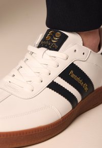 Vita lädersneaker med marinblå ränder och guldtryck. Har en strukturerad gummisula och vita snören, samt en vadderad ankelskaft.