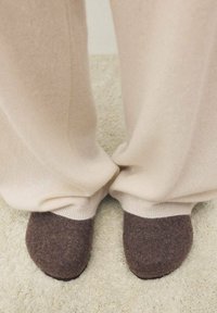 Pantofole di feltro marroni con punta arrotondata e un risvolto di colore più chiaro, indossate con pantaloni beige larghi su una superficie chiara e texturizzata.