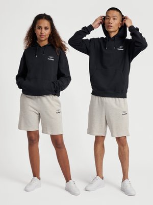 Dwie osoby ubrane w czarne bluzy z kapturem i jasnoszare szorty z trampkami, stojące na jednolitym białym tle.