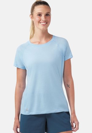 Donna sorridente, indossa una camicia azzurro chiaro a maniche corte e pantaloncini blu scuro, con i capelli legati e un piccolo tatuaggio di un uccello sull'avambraccio sinistro.