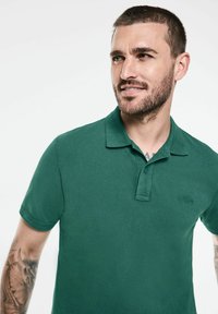 Zelená polokošile vyrobená z bavlny, s límcem, krátkým rukávem a decentním logem na hrudi. Hladká textura a klasický střih.