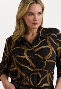 Svart bluse med et mønster av gullkjeder, med lange ermer, krage og en glatt tekstur, som fremhever et elegant design.
