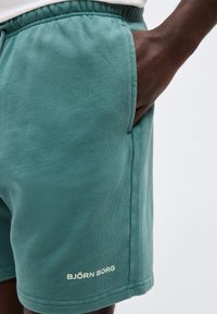 Κοντινό πλάνο ενός ατόμου που φοράει τιρκουάζ σορτς Björn Borg με το ένα χέρι στην πλαϊνή τσέπη σε απλό φόντο.