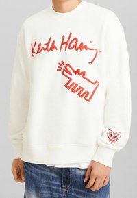 Vit tröja med röd text och grafik, med "Keith Haring" signatur, abstrakt design och hjärtillustration på ärmen.