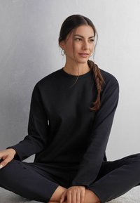 Schwarzer, langärmeliger Rundhalsausschnitt-Sweatshirt mit strukturiertem Finish, kombiniert mit passenden Hosen. Modell sitzt mit übergeschlagenen Beinen vor einem sanften, neutralen Hintergrund.