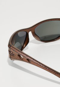 Óculos de sol castanhos com uma armação texturizada, lentes cinzas e detalhes recortados nas hastes. Design leve, acabamento suave.