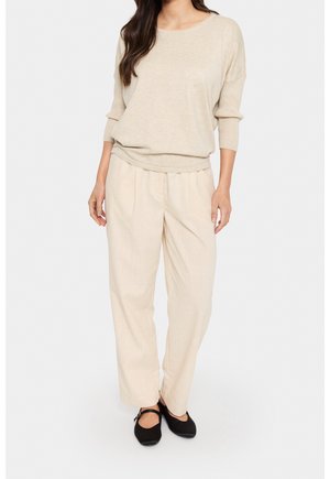 Beżowy sweter dzianinowy z rękawami 3/4, zestawiony z jasnobeżowymi, luźnymi spodniami i czarnymi płaskimi butami. Gładka tekstura, casualowy design.