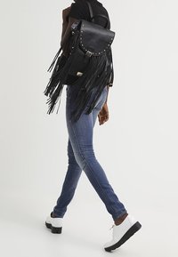 Mochila de cuero negra con detalles de flecos, acentos de tachuelas y cierre de solapa. La modelo lleva puestos unos pantalones vaqueros azules y zapatos blancos, alejándose de la cámara.