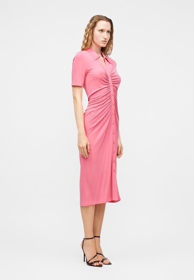 Γυναίκα που φοράει ροζ midi φόρεμα εφαρμοστό, με ρυτίδες και κουμπιά στο μπροστινό μέρος, με κοντά μανίκια, συνδυασμένο με μαύρα πέδιλα με λουράκια και ψηλό τακούνι.