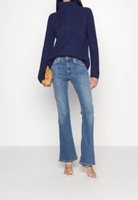 Marinblå ribbad polotröja, ljusblå utsvängda jeans, sandaler med genomskinlig klack och en tan-färgad väska med rynkad design.