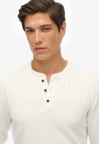 Chemise à manches longues crème avec un col henley, trois boutons marron et une texture lisse. Présente une coupe ajustée et un design minimaliste.