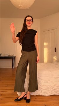 Νεαρή γυναίκα με μαύρο τοπ και ελιά φαρδιά παντελόνια χαιρετάει μέσα σε ένα απαλά φωτισμένο υπνοδωμάτιο με ξύλινο πάτωμα.