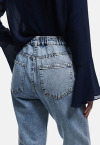 Jeans de mezclilla azul claro con cintura elástica, que cuentan con dos bolsillos traseros y un ajuste ligeramente holgado. Combinando con una camisa azul oscuro.