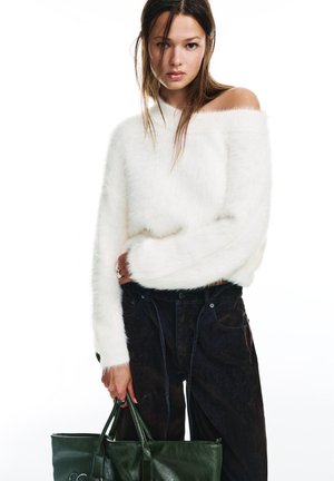 Mladá žena v bielom chlpatom svetri s odhalenými ramenami a čiernych nohaviciach, drží veľkú tmavozelenú koženú tašku po boku.