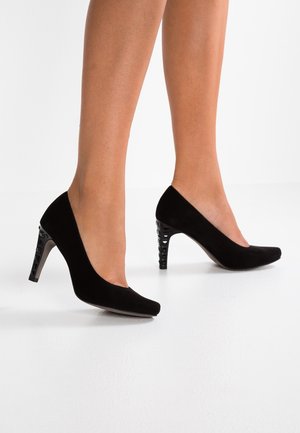 Benen met zwarte suède pumps met getextureerde blokhakken, staand op een witte vloer tegen een witte achtergrond.