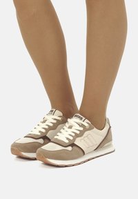 Sneakers beige e crema realizzati in camoscio e mesh, con lacci bianchi, accenti marroni e suola in gomma. Mostrano un logo sulla linguetta.