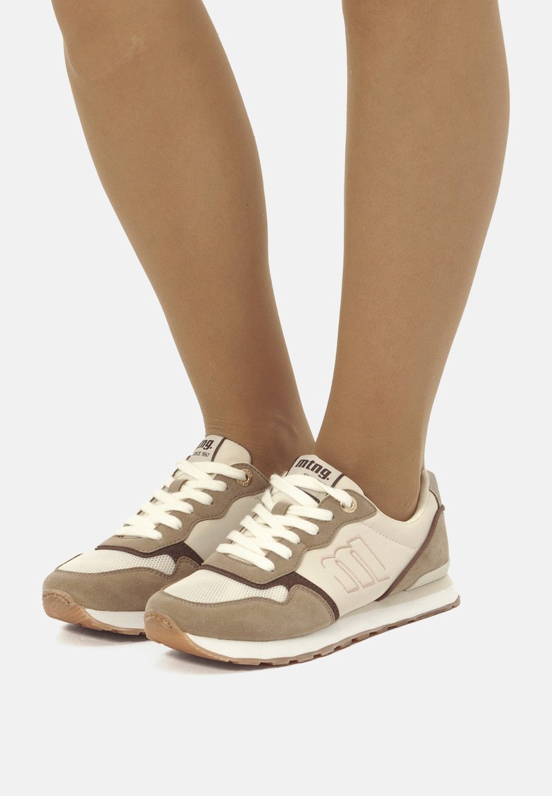 Sneakers beige e crema realizzati in camoscio e mesh, con lacci bianchi, accenti marroni e suola in gomma. Mostrano un logo sulla linguetta.