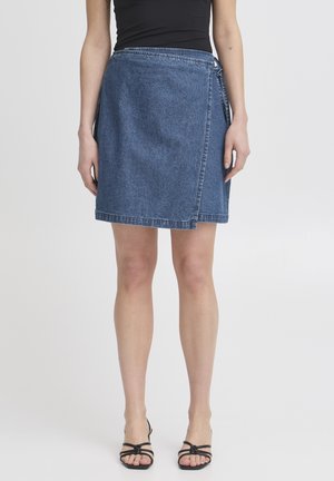 Vrouw draagt een blauwe denim wikkelrok, een zwart mouwloos topje en zwarte sandalen met bandjes, staand tegen een effen lichte achtergrond.