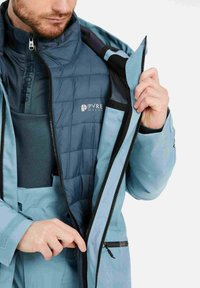 Blauwe waterdichte jas met een gewatteerde binnenlaag. Voorzien van een hoge kraag, ritszakken en contrasterende zwarte ritsdetails.