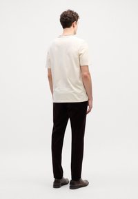 T-shirt crème à manches courtes avec un col rond, associé à un pantalon sombre. Le modèle se tient devant un fond neutre. Design minimaliste.