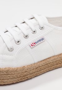 Sneaker bianco in tela con suola spessa in juta intrecciata, lacci bianchi infilati attraverso occhielli in metallo e un'etichetta del marchio in tessuto sul lato.