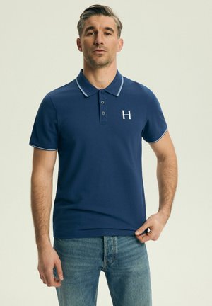 Mann trägt ein dunkelblaues Polohemd mit weißem Rand und einem "H"-Logo auf der Brust, kombiniert mit blauen Jeans, stehend vor einem einfarbigen Hintergrund.