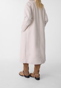 Donna che indossa un lungo cappotto beige con le mani nelle tasche, abbinato a stivali in cammello di camoscio fino a metà polpaccio con fibbie, in piedi su uno sfondo bianco.
