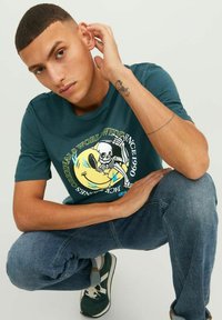 Camiseta de algodón color teal con un diseño gráfico de un esqueleto y una cara sonriente. Combinada con jeans de mezclilla azul y zapatillas verdes.