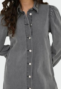 Chemise grise à boutonner en denim avec un col, des manches bouffantes et des boutons argentés sur le devant, présentant des détails de couture contrastants.