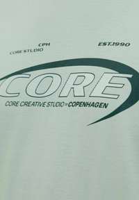 Svijetlozelena pamučna majica s odvažnim crno-bijelim grafičkim uzorkom na kojem se nalazi tekst "CORE" i dodatni manji tekstualni elementi.