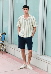 Beige short-sleeve shirt with blue vertical stripes, paired with navy shorts and light beige sneakers; standing against glass background.