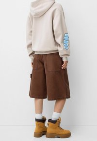 Beige huvtröja med blå grafik på ärmen, bruna baggy shorts och gula snörskor. Vita ankelsockor synliga ovanför stövelkanterna.