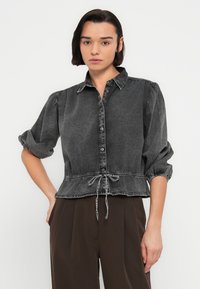 Μπλούζα από μαύρο τζιν με κουμπιά στο εμπρός μέρος, φουσκωτά μανίκια και σφιχτή μέση με κορδόνι. Ελαφρώς ξεθωριασμένη υφή.