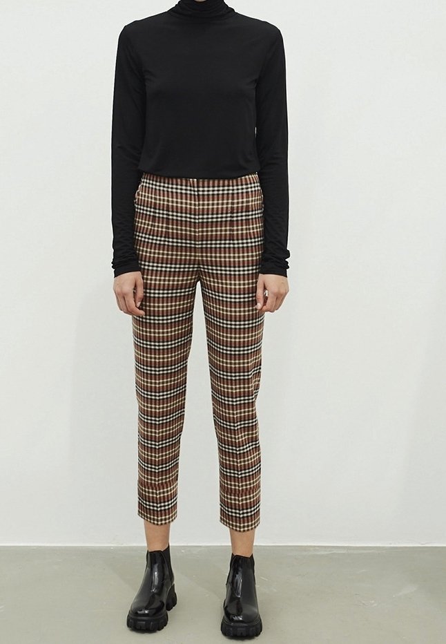Personne portant un haut noir manches longues, un pantalon court marron et blanc à carreaux, et des bottines noires épaisses, debout contre un mur uni.