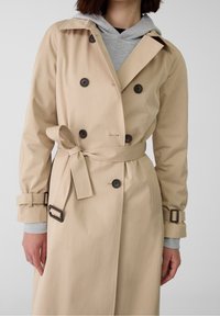 Beige trench coat with double-breasted design, large dark buttons, belted waist, and prominent collar. Worn over a gray hoodie.