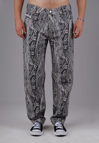 Man wearing black and white patterned pants and black sneakers with white laces, standing with arms relaxed by sides against gray background.