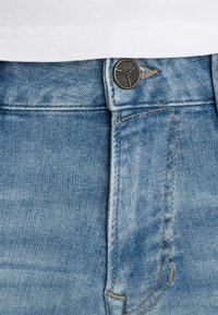 Kadar u blizini plavih traperica s pojasom i metalnom kopčom označenom "PME Legend Jeans" te dijelom bijele košulje iznad.