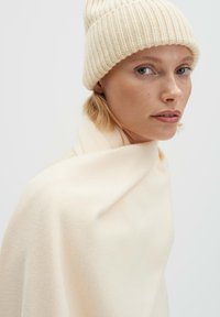 Beige wollen poncho valt over de schouders, gecombineerd met een licht crèmekleurige, geribbelde gebreide muts. Gladde, zachte textuur, minimalistisch ontwerp.