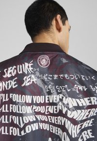 Chaqueta negra y granate con un diseño con cuello. Presenta patrones de texto en blanco y en varios idiomas, con el logo del Manchester City en la parte posterior.