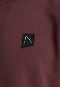 Sudadera de color burdeos con un acabado texturizado, presenta un pequeño parche negro en forma de triángulo con un diseño gráfico blanco.