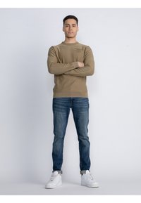 Hellbraunes langärmliges Sweatshirt mit Rundhalsausschnitt, kombiniert mit blauen Jeans und weißen Sneakers. Das Sweatshirt hat dezente Markenkennzeichnungen auf der Brust.