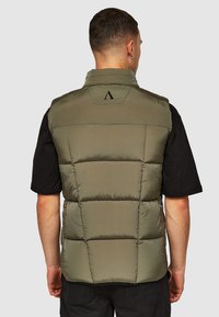 Groene donzen vest met horizontaal gestikte delen, hoge kraag en logo-detail op de achterkant. Draagt over een zwart T-shirt met korte mouwen.