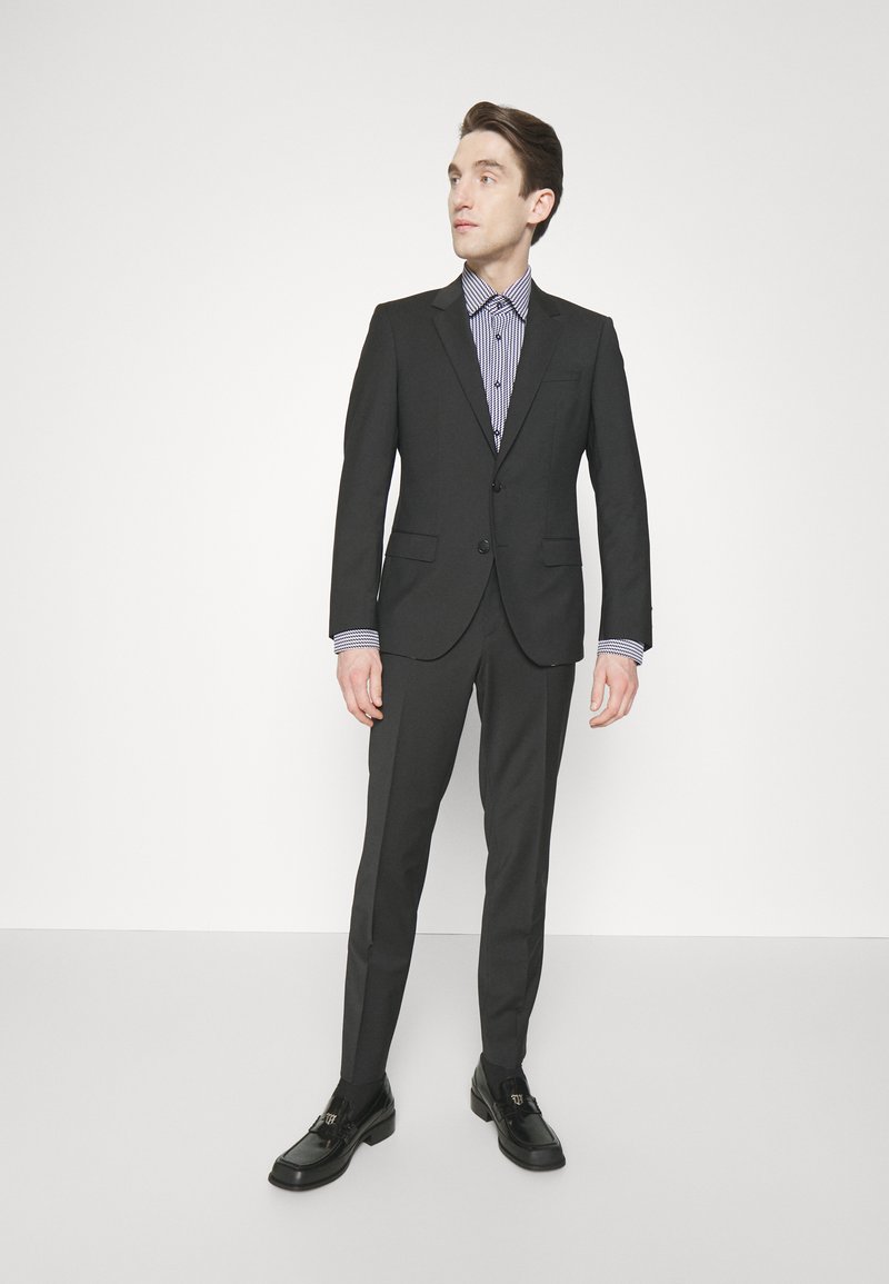 Traje negro con un corte ajustado, camisa blanca con patrón de cuadros en navy y mocasines negros. Tela suave, hombros estructurados y pantalones ajustados.