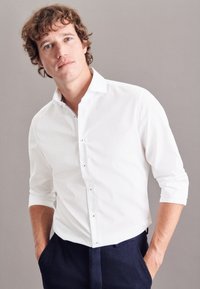 Camicia bianca con bottoni, maniche lunghe arrotolate fino ai gomiti, dotata di piccoli bottoni metallici, texture liscia e un leggero splendore.