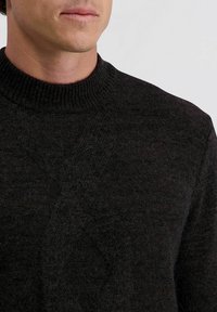 Donker, gebreid vest met een hoge, geribbelde kraag. Heeft een textuur met een ruitpatroon aan de voorkant. De stof lijkt zacht en warm.