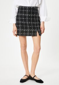 Mini-jupe à carreaux noirs et blancs en tissu texturé, avec une fente sur le côté. Portée avec des chaussures plates noires et une chemise blanche.