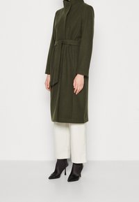 Olive green belted coat with a high collar, long sleeves, straight cut, paired with cream wide-leg trousers and black heeled boots.