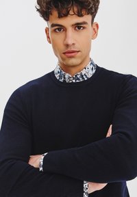 Granatowy sweter dzianinowy z teksturowanej tkaniny i wzorzystym białym kołnierzem w niebieskie kwiatowe wzory. Model ma skrzyżowane ramiona.